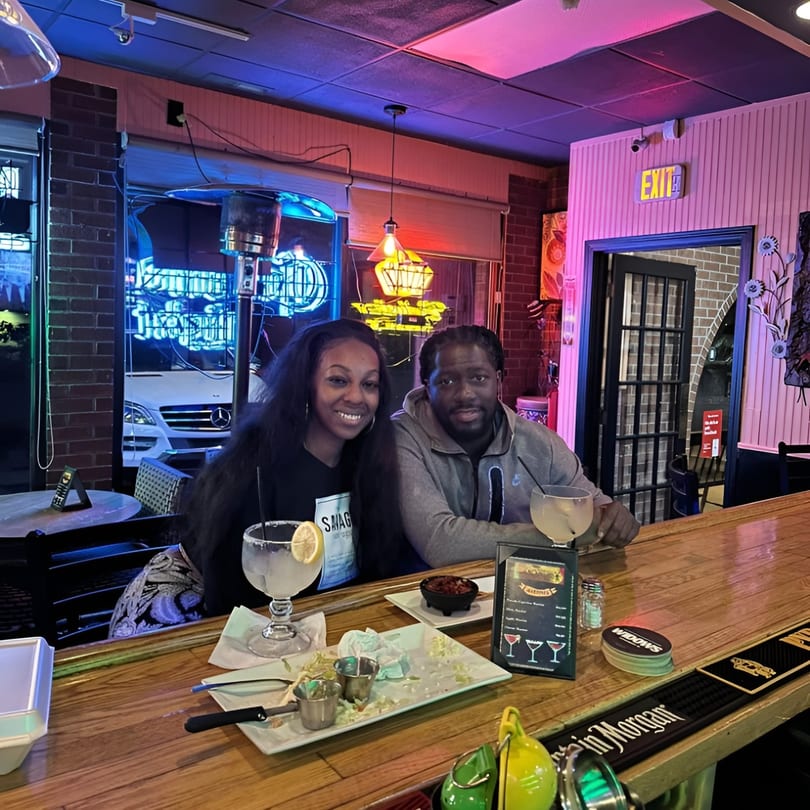 A couple smiles at the camera while sitting at a bar with drinks and food at El Don Quijote.