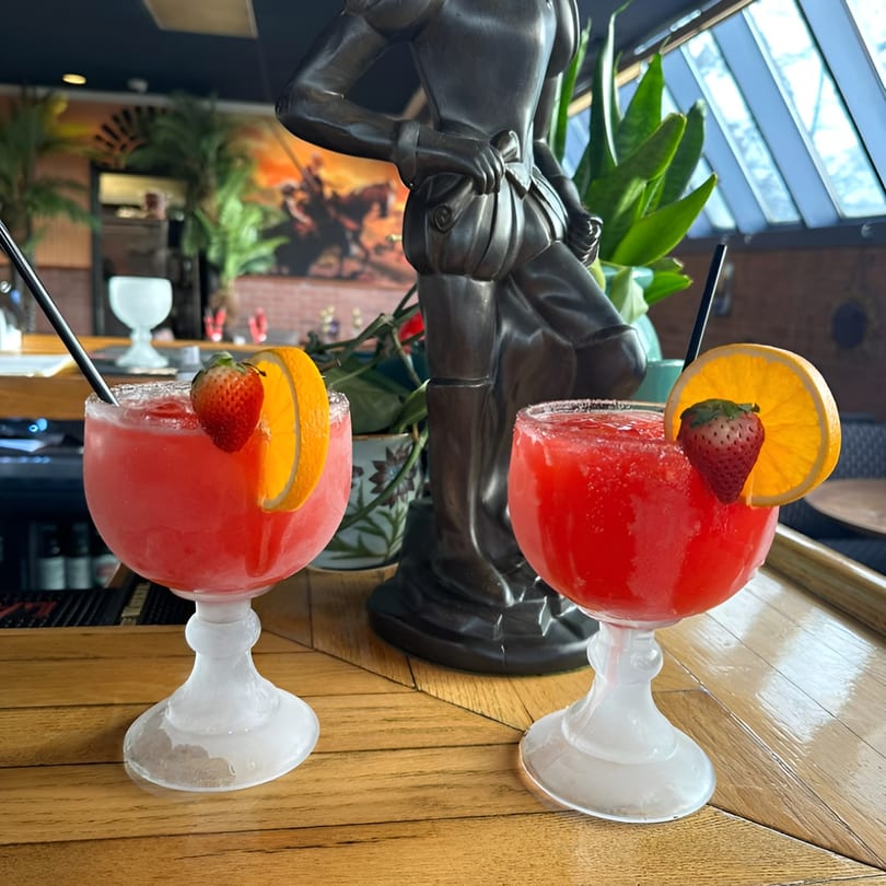 Two strawberry margaritas with orange slices and strawberries on a wooden bar top.