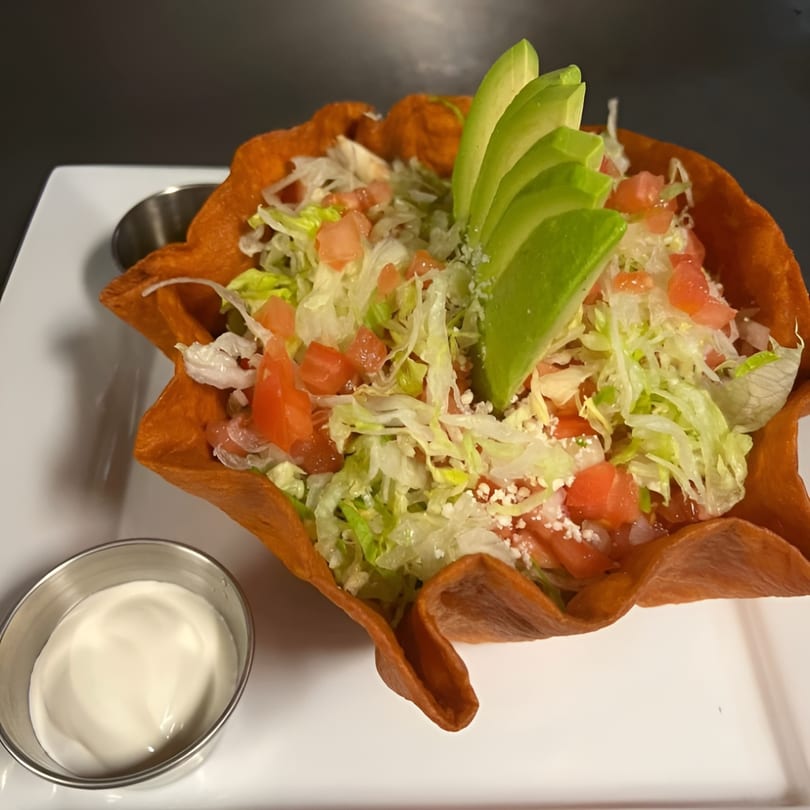 A vibrant taco salad in a crispy orange tortilla bowl, topped with lettuce, tomatoes, cheese, and avocado slices.
