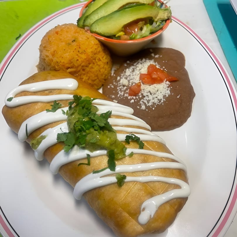 A chimichanga drizzled with sour cream and guacamole, served with rice, refried beans, and avocado salad.