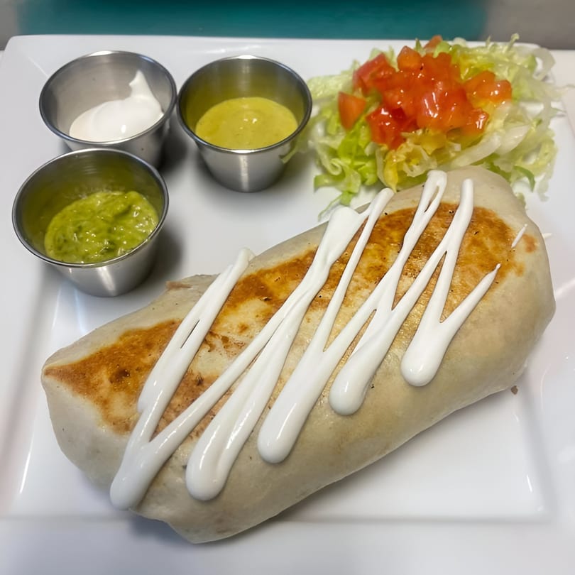 A large, grilled burrito drizzled with white sauce, served with guacamole, sour cream, and salsa.